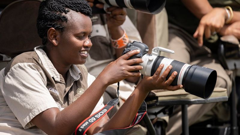 A young woman in brown and beige Canon-branded uniform holds a Canon camera and smiles as she looks at the rear screen.