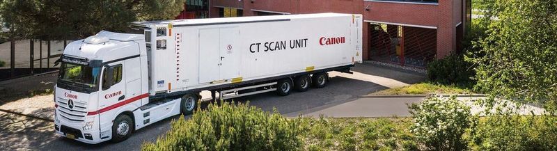 A Canon branded lorry parks a mobile CT scanning unit, with CT SCAN UNIT and the Canon logo printed on the side. It is outside what appears to be a brown brick office building, surrounded by trees and bushes.