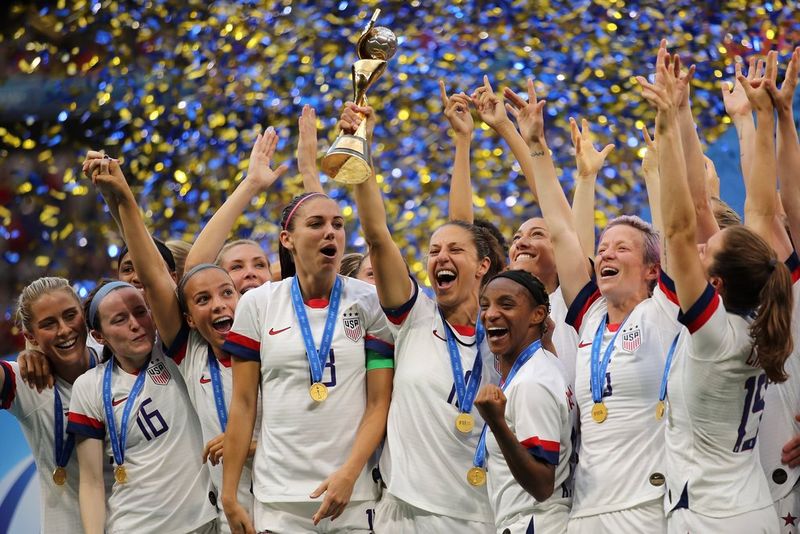 Carli Lloyd, captain of the US women's national football team, lifts a glittering gold trophy above her head as her teammates celebrate in a shower of confetti.