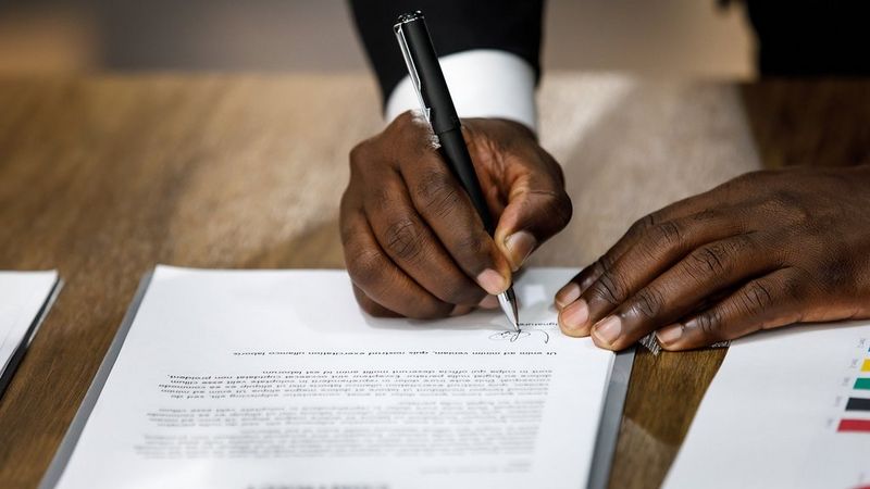 A pair of hands on a wooden desk, signing what appears to be a letter with an elegant black pen.