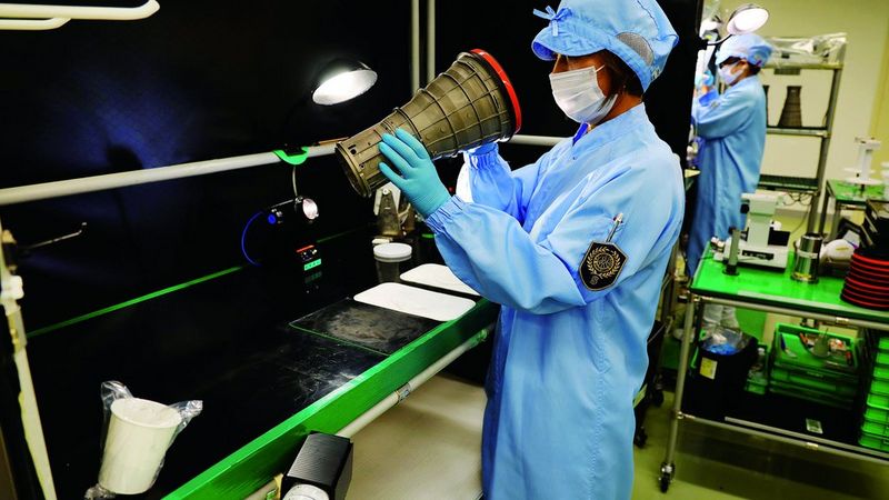 Un operaio con tuta blu, copricapo e guanti ispeziona un oggetto a forma di obiettivo tenendolo in mano.