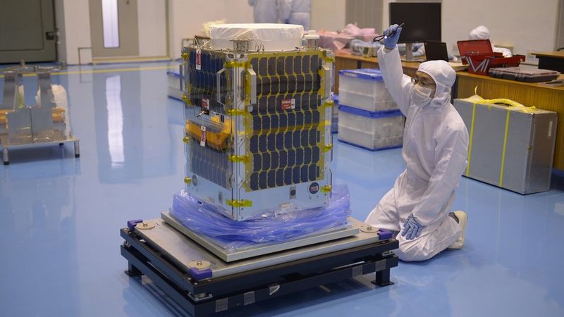 Una persona in tuta bianca, maschera e guanti in lattice blu è inginocchiata davanti a un microsatellite Canon. Ha un braccio sollevato e impugna una torcia mentre ispeziona il telaio della struttura.