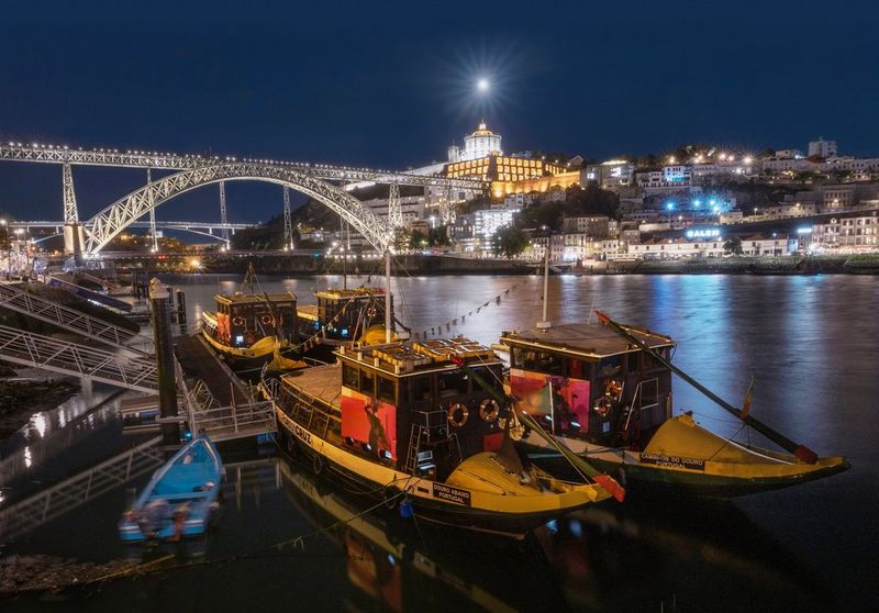 The Moon glows brightly in the dark sky, above a night scene of a river, bridge and boats with a cityscape behind. Taken on a Canon EOS R8.