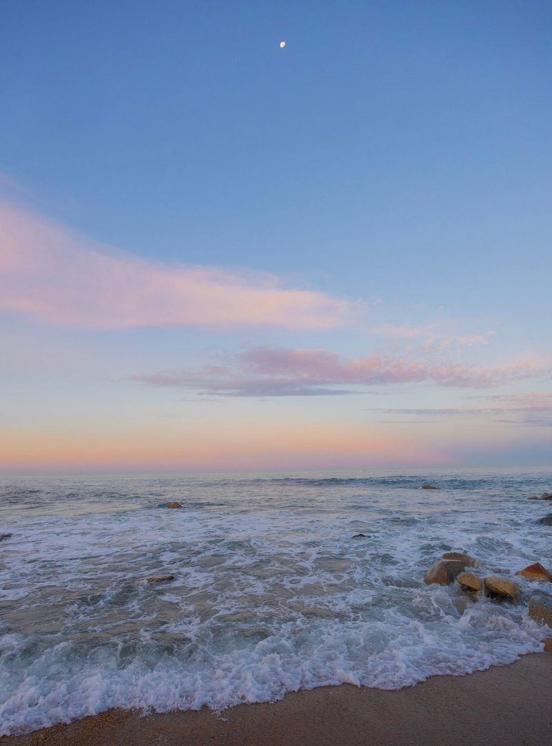 The Moon visible high up in the sky above light orange clouds and a body of water, taken at sunrise on a Canon EOS R8.