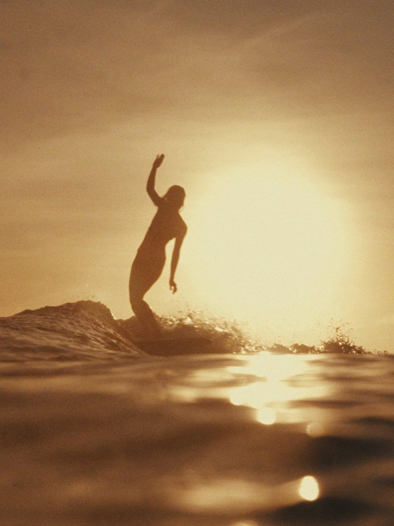 Image d’une femme entrain de surfer avec un coucher de soleil dans le dos