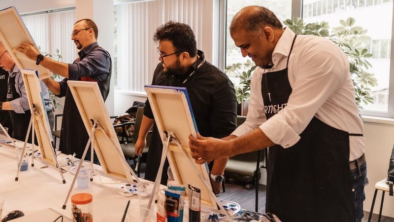 Drie mannen staan naast elkaar op doeken te schilderen. Hun schildersezels staan op tafels voor hen.