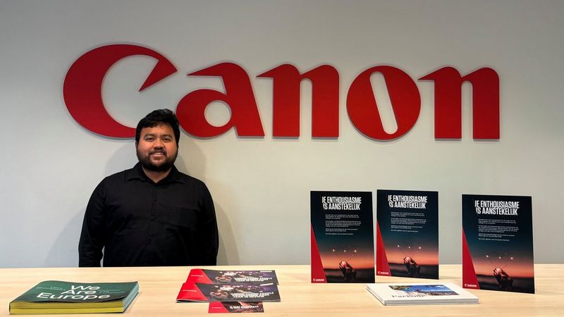 Muhammad Rezqi staat links voor een groot rood Canon-logo. Voor hem staat een tafel met brochures en folders.