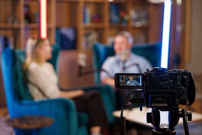 A camera recording an interview between two people seated in a studio, with the subjects softly blurred in the background.