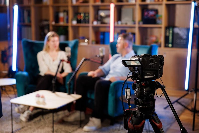 Two blurred people in armchairs, talking to each other. In the forefront of the image is a Canon camera on a tripod, filming them as they talk.