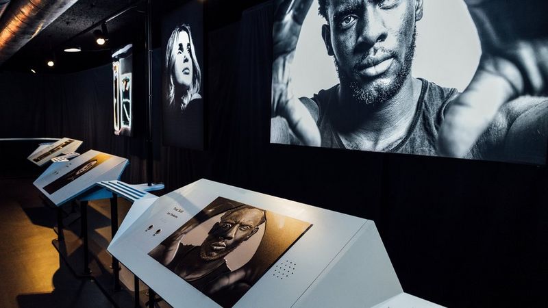 A dimly lit, modern gallery featuring large black and white portraits mounted on dark walls. In the foreground is a photograph of a man looking directly at the camera with his hands raised near his face. Further back, another shows a blonde woman with shoulder length hair looking upward and to the side. Pedestals stand in front of the portraits, showing smaller versions of the artwork, descriptive text and buttons for an interactive experience.