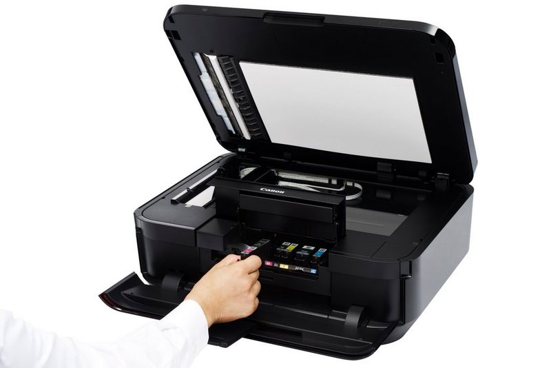 A user's hand removes one single-colour ink cartridge from a Canon multi-function device with its scanner lid and access flap open.