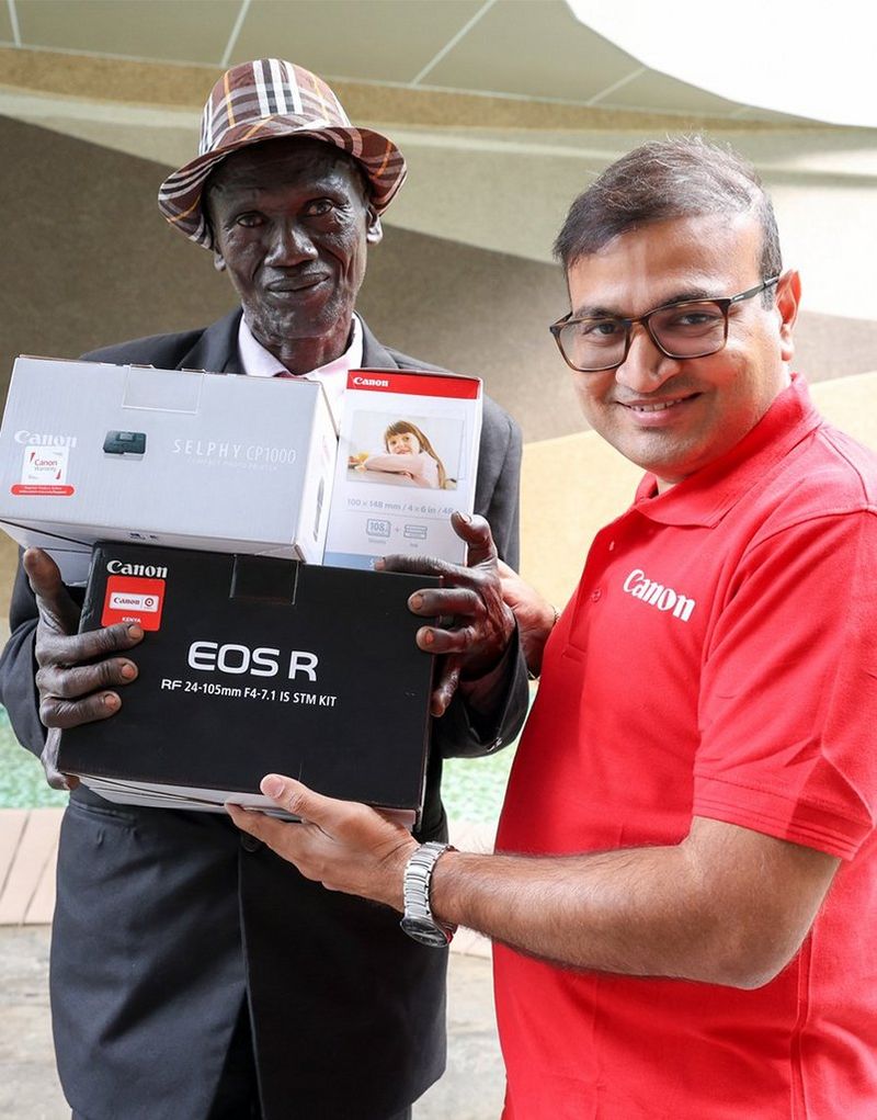 Two men stand side by side. One is wearing a red Canon polo shirt and handing the other a selection of Canon products.