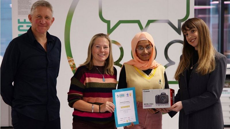 Four people stand side by side: a man, dressed in black. A woman in a striped sweater, holding a certificate. A bespectacled young woman in a peach headscarf and yellow sweater, and a long-haired young woman in a black business suit. She holds a box for a Canon EOS 250D camera.