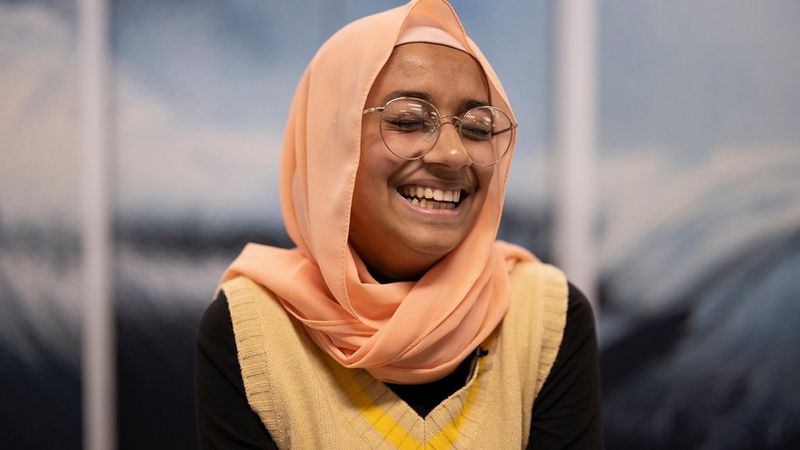A young bespectacled woman in a peach-coloured headscarf and glasses laughs with her eyes scrunched shut.