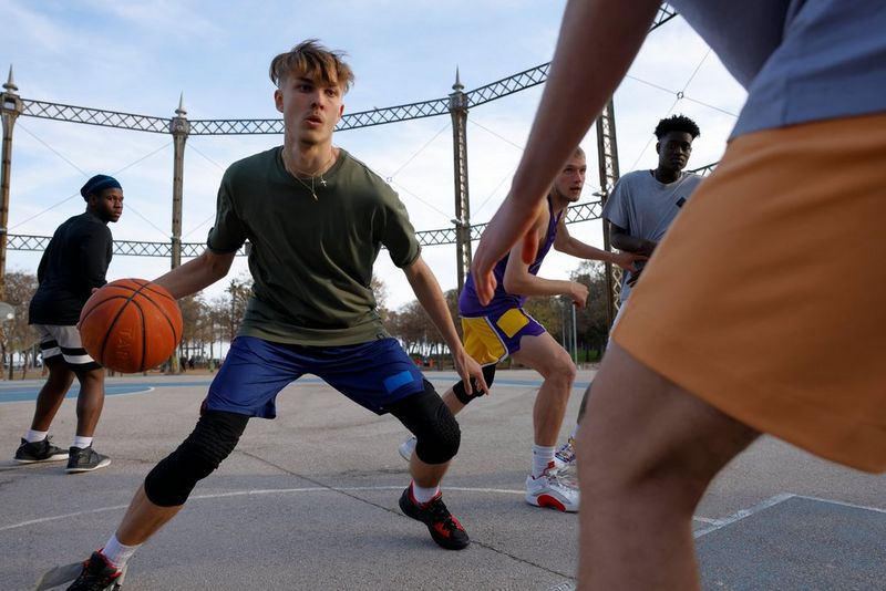 A young man is poised, legs apart, on a sports court, orange basketball in one hand. Several more players can be seen around him.