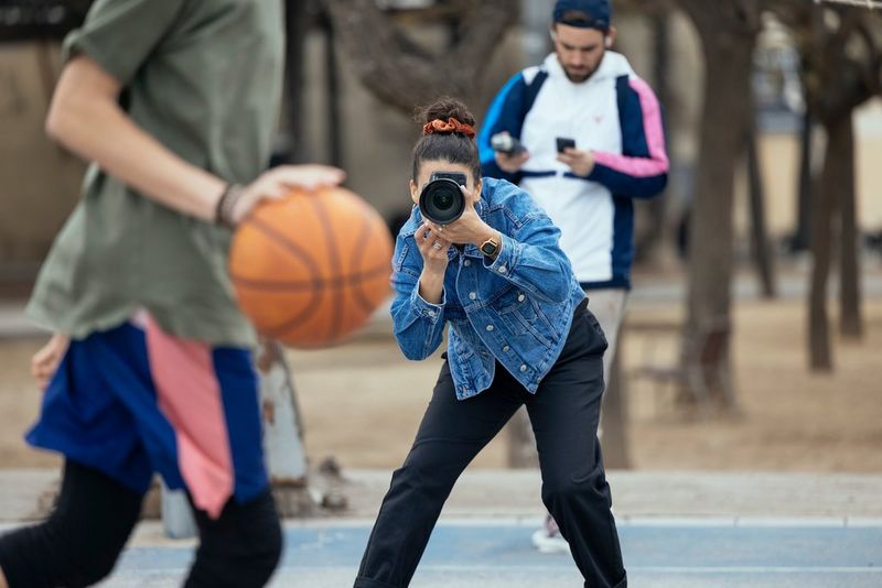 A female photographer leans over slightly to take a picture of a streetball player on a hard court as they are about to bounce the ball.