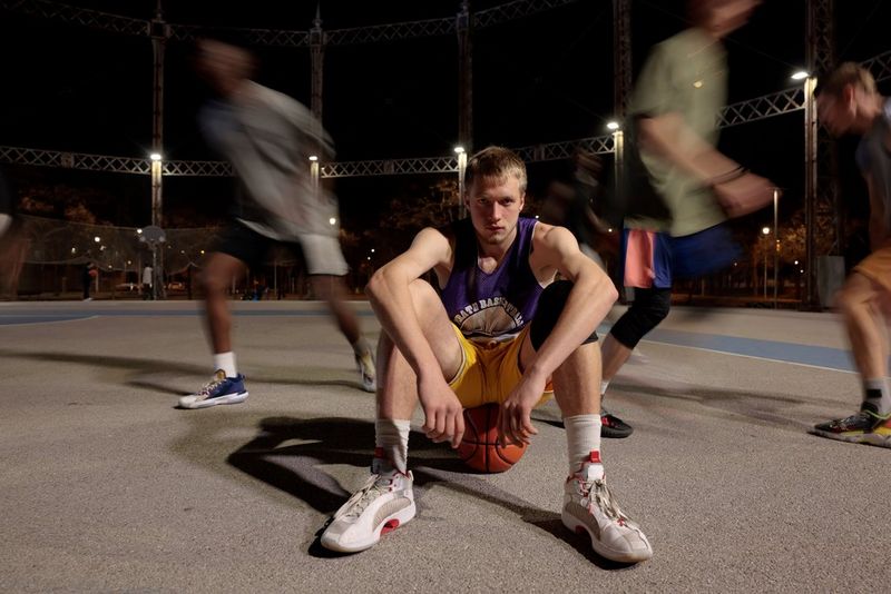 A young man sits balanced on an orange ball on the floor of a sports court, his legs drawn up towards him. Around him you can see several other players, blurred as they move.
