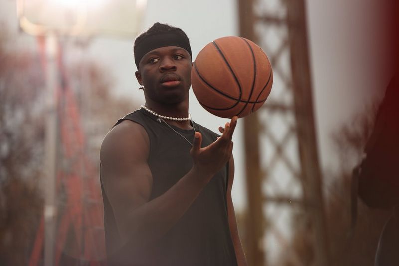 A man wearing a sleeveless black top and matching bandana spins a basketball casually on the middle finger of his right hand.