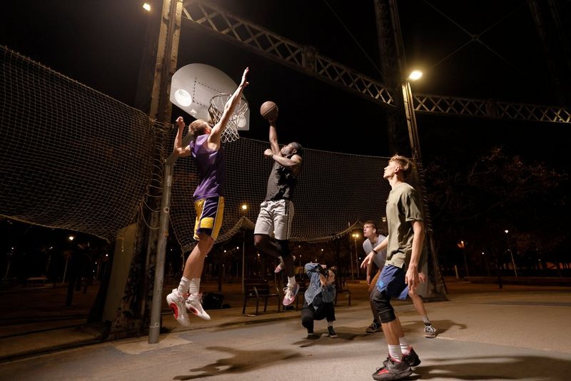 A woman crouches down to take a photograph of two men jumping up at a streetball hoop. The court is dark and lit by dim floodlights.