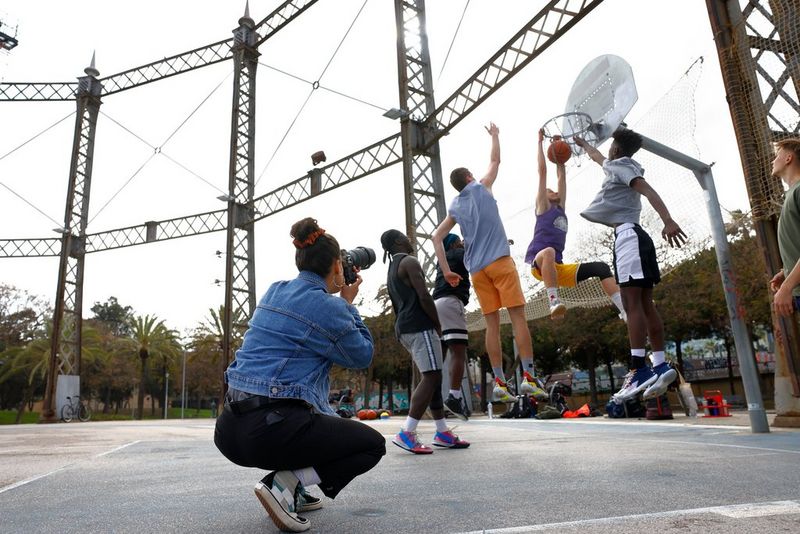 A woman crouches down to take a photograph of a group of men jumping up at a streetball hoop.