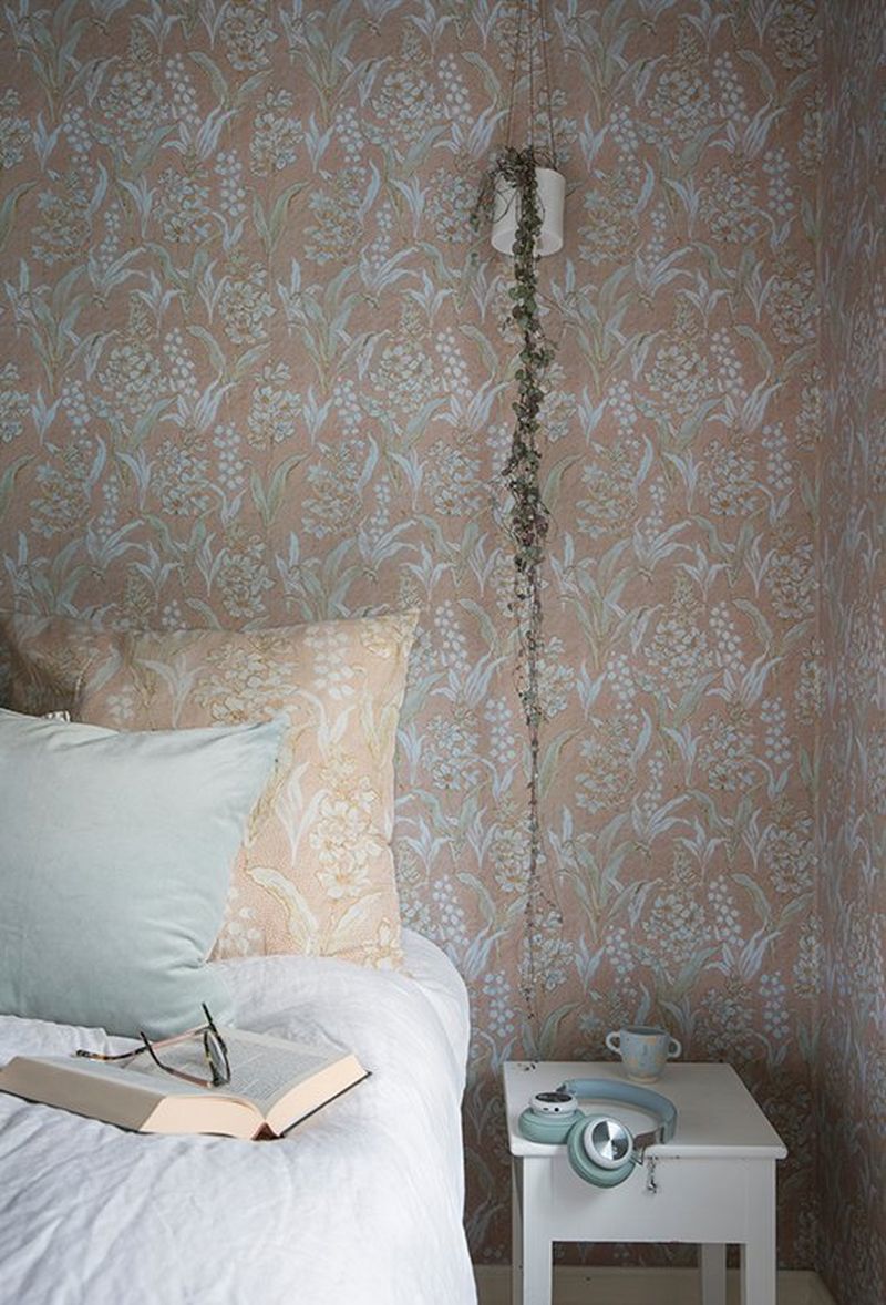 A partial view of a bed with a bedside table, set against the reproduction wallpaper with Lily of the Valley pattern.