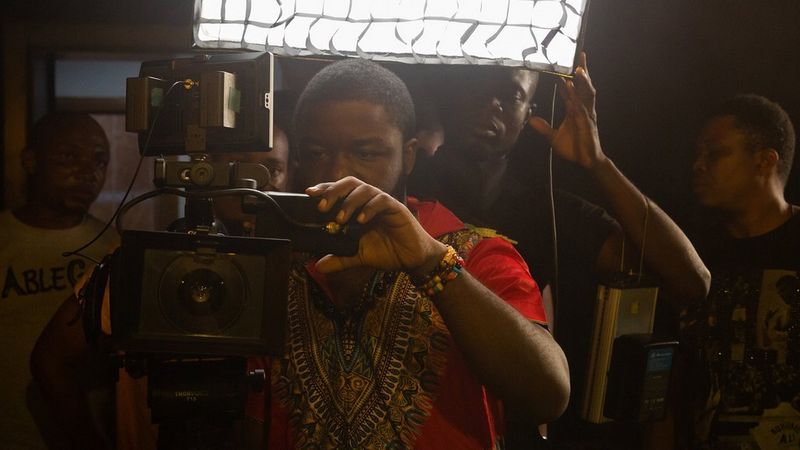 A man wearing a red t-shirt is holding and looking through a Canon camera in Lagos, Nigeria. Behind him, two men look in the same direction.