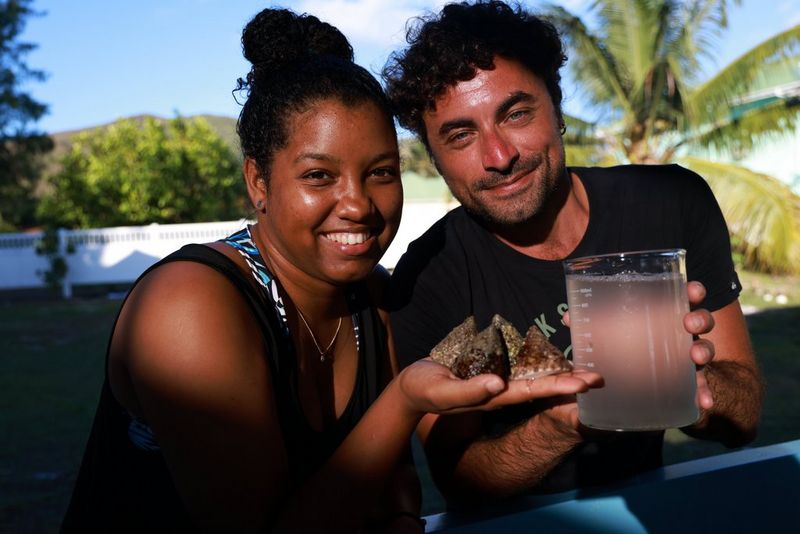 Priya (à gauche) sourit en tenant dans la paume de sa main quatre escargots de forme conique, appelés trochus. À côté d'elle, le Dr Luca Saponari, souriant, tient un bécher en borosilicate contenant des gamètes de corail.