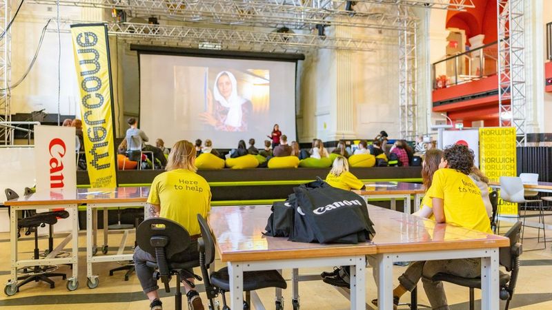 Students in the huge, high-ceiling hall of 20 Bank Plain. Most are on beanbags and two at the back are sat at a table wearing t-shirts with ‘here to help’ printed on the back. They are all watching a huge projector screen showing a woman in a white headscarf.