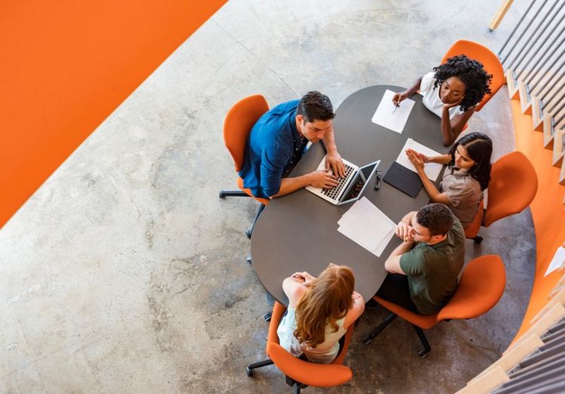 Overhead shot of office workers sat round an oval-shaped table on orange chairs, spread out with papers and a laptop computer.