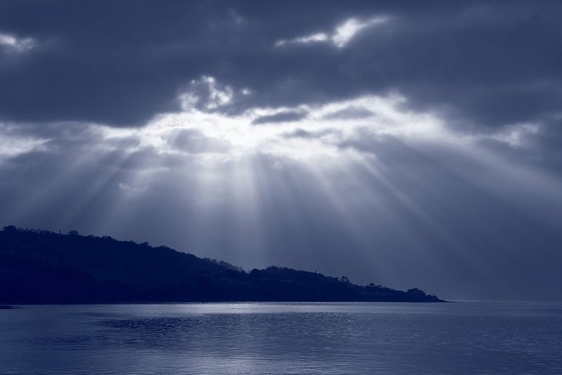 Un paesaggio marino monocromatico dai toni blu, con i raggi del sole che splendono attraverso le nuvole scure sopra la silhouette di una costa collinare.