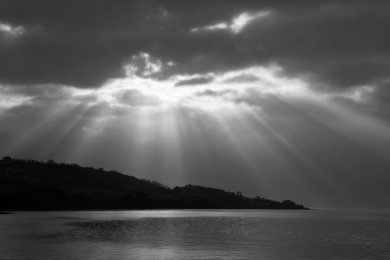 Un paesaggio marino in bianco e nero, con i raggi del sole che splendono attraverso le nuvole scure sopra la silhouette di una costa collinare.
