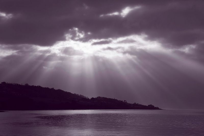 Un paesaggio marino monocromatico dai toni violacei, con i raggi del sole che splendono attraverso le nuvole scure sopra la silhouette di una costa collinare.