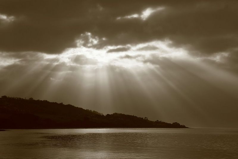 Un paesaggio marino monocromatico dai toni seppia, con i raggi del sole che splendono attraverso le nuvole scure sopra la silhouette di una costa collinare.