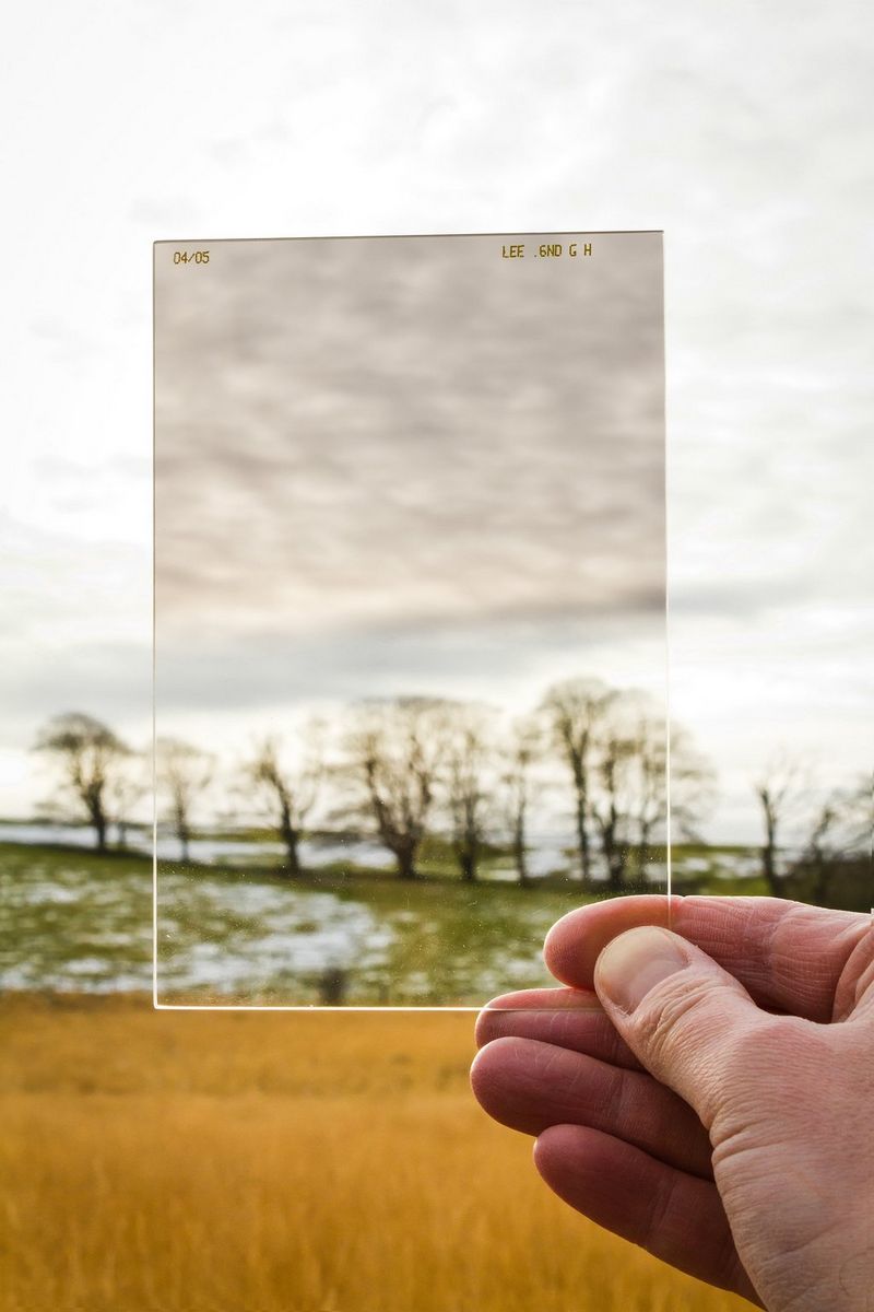 A hand holding ND filter against a landscape with trees and cloudy sky. 