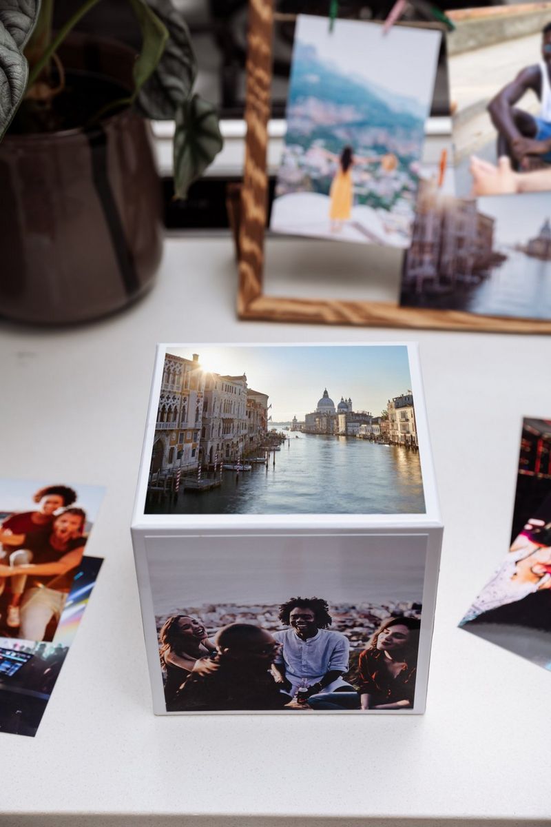 A Canon Photo Cube on a table, with a picture of Venice on the top and a group of people on the side. Other photo prints are visible in the background. 