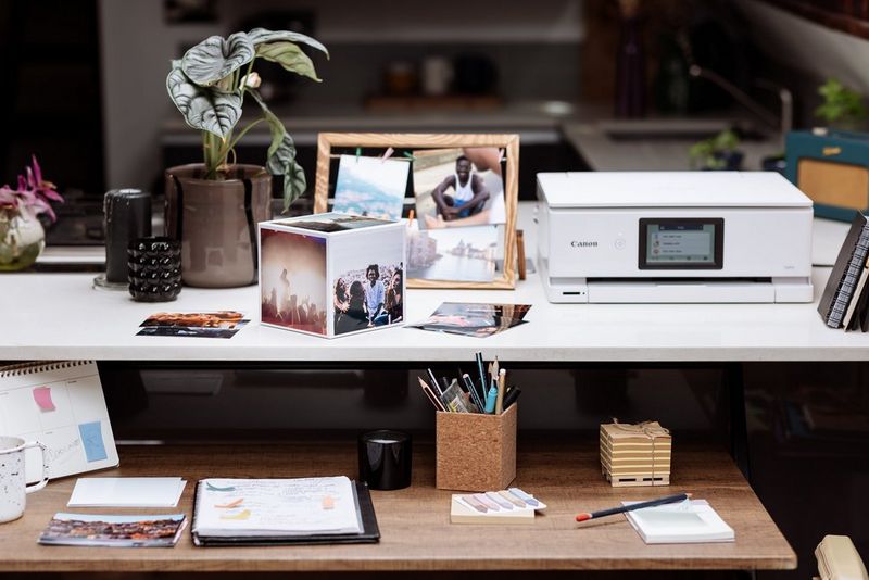A Canon Photo Cube sits on a desk, a Canon printer, plants, photo frames and other trinkets next to it. 