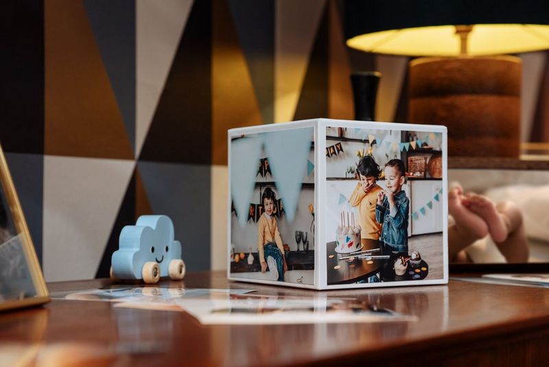 A Canon Photo Cube with children's party pictures around the sides sits on a surface next to other photos and trinkets.