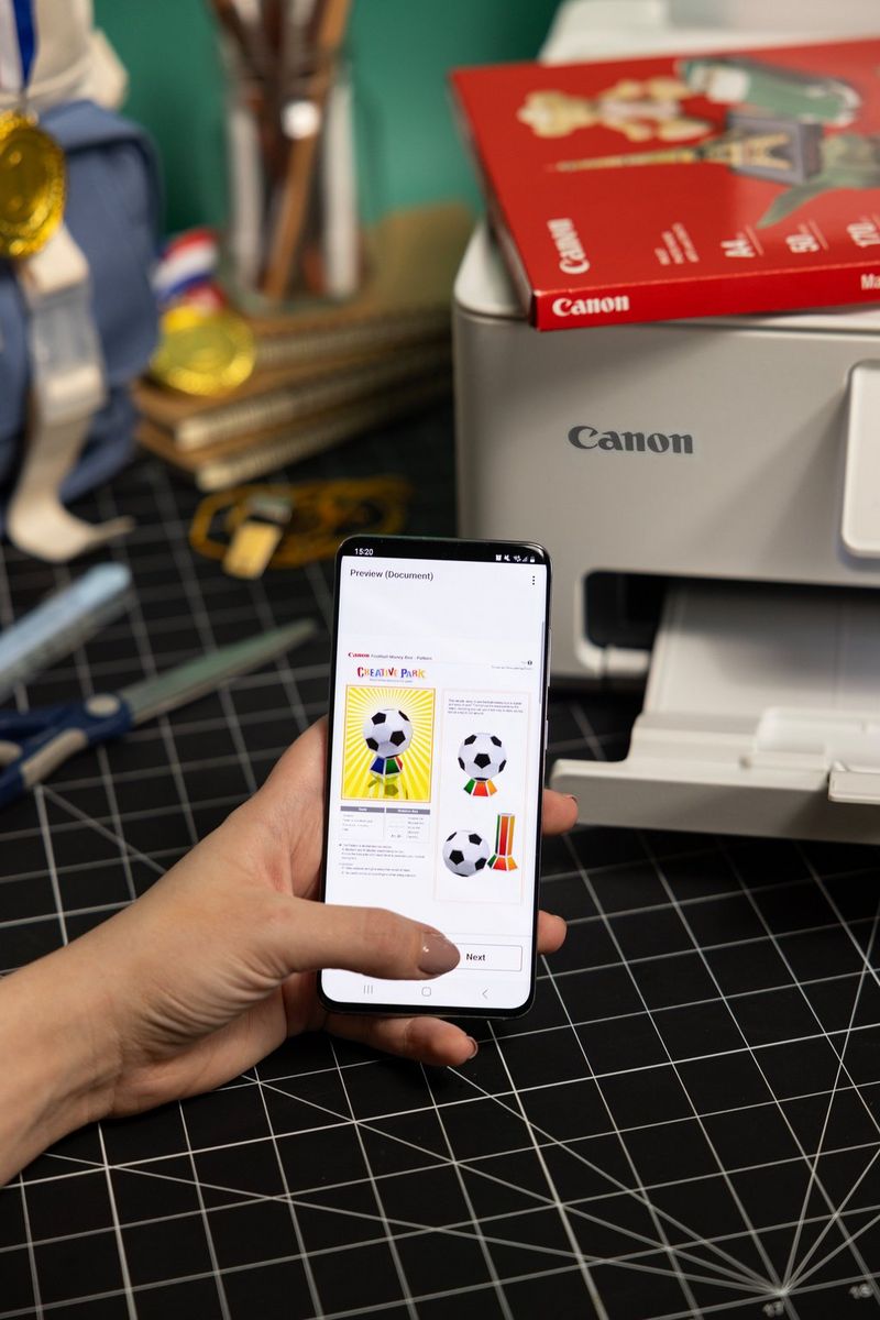 Een smartphone met daarop Canon Creative Park-sjablonen in voetbalthema naast een Canon PIXMA-printer op een snijmat