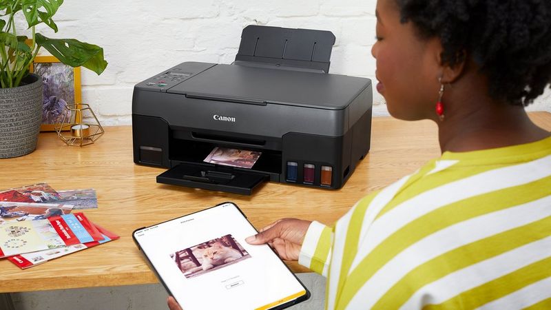 A Canon printer sits on a wooden desk, mid-printout. A woman sits in front of it with some packs of printer paper, looking at an image on a mobile tablet.