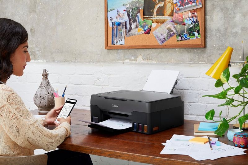 A young woman holding a smartphone sits at a desk with a Canon PIXMA printer on it.