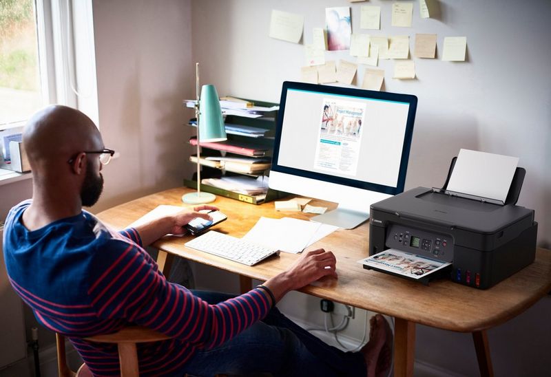 A person sits working at a wooden desk with a monitor, keyboard and Canon printer on it. Stuck on the wall above the desk are various small sheets of paper with notes written on them. 