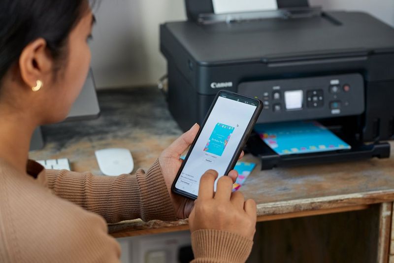 Hands holding a smartphone displaying a Canon print app. The image on the smartphone screen is emerging from the printer in the background.