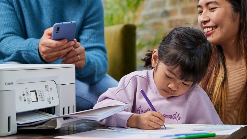 A child sits on a smiling woman's knee colouring in printouts from a Canon PIXMA printer. Hands holding a smartphone can be seen in the background.