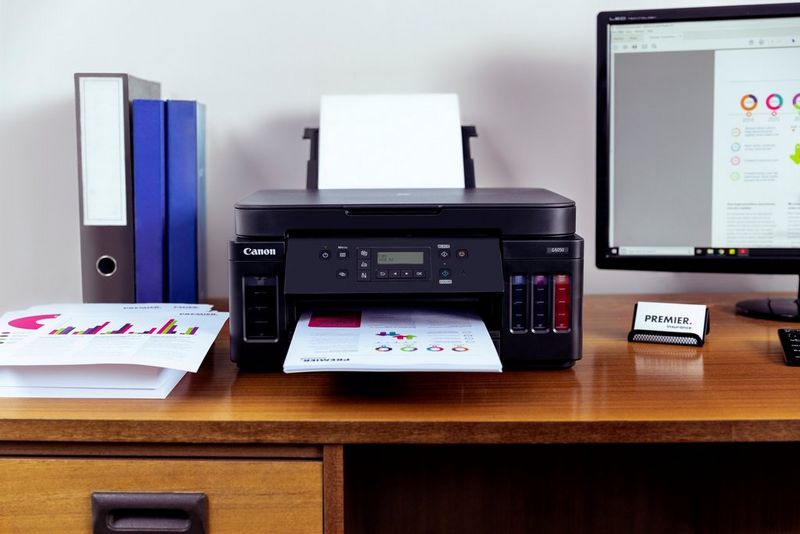 Canon printer on desk, with paper in both input and output trays. Lever arch files and stacked paper to the left and a computer screen to the right. Shot on Canon.