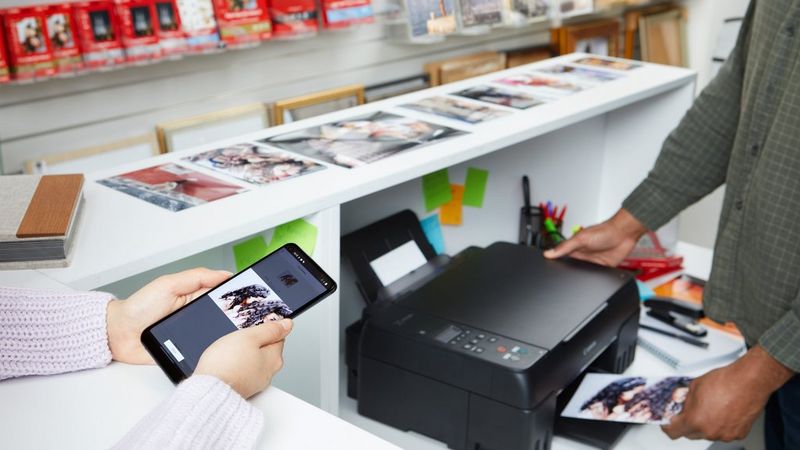 Een tablet met de geopende Canon PRINT-app ligt op een Canon-printer waar een foto uitkomt, en er liggen andere foto's naast op een bureau.