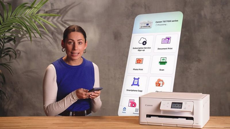 En person, der holder en smartphone, sidder bag et skrivebord med en Canon PIXMA-printer, og en visning på telefonskærmen bagved viser Canon PRINT-appen.
