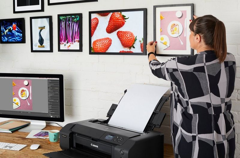 Eine Frau hängt ein gerahmtes Bild an einer Wand auf. Auf ihrem Schreibtisch befinden sich ein Computer und ein Canon PIXMA PRO-200 Drucker.