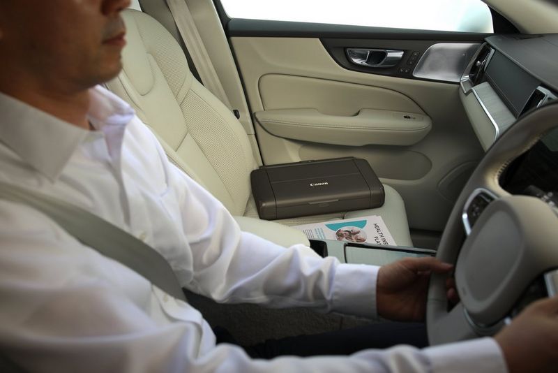 A man drives his car with a PIXMA TR150 on the passenger seat beside him. 