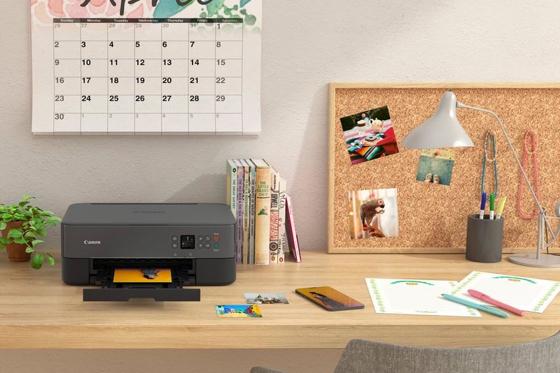 Una stampante Canon PIXMA su una scrivania in legno accanto a una bacheca, libri e articoli di cancelleria.