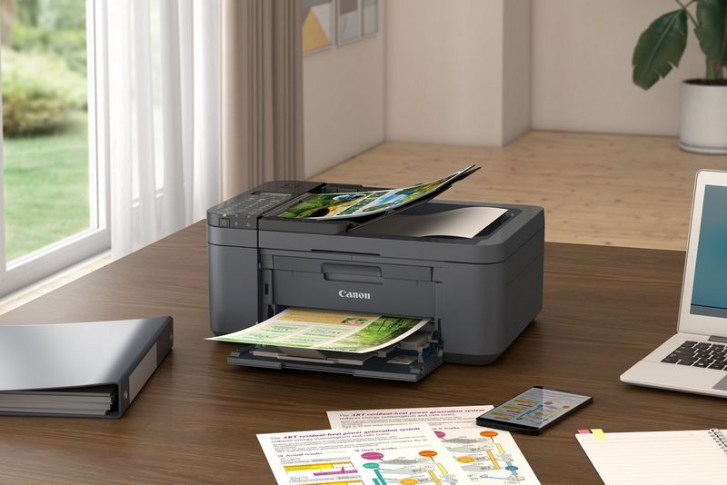 A printer sits on a table with printouts in the output trays, in a home setting.